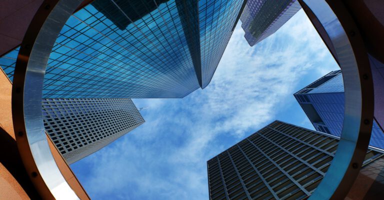 Upward view of skyscrapers framed by a circular structure, with glass facades reflecting a blue sky.