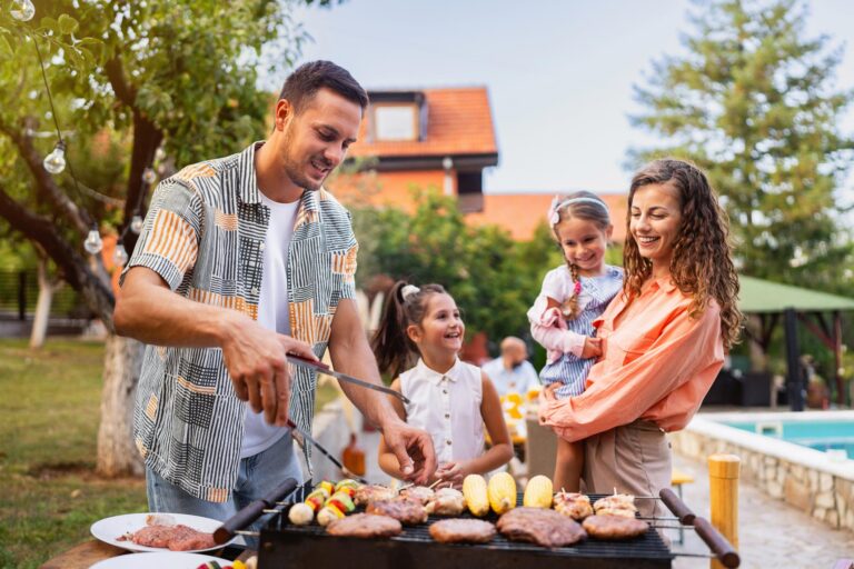 Smiling man grills meat and vegetables while a woman and two young girls enjoy time together outdoors.